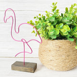Pink flamingo decoration on a wooden base next to a potted plant with green leaves.