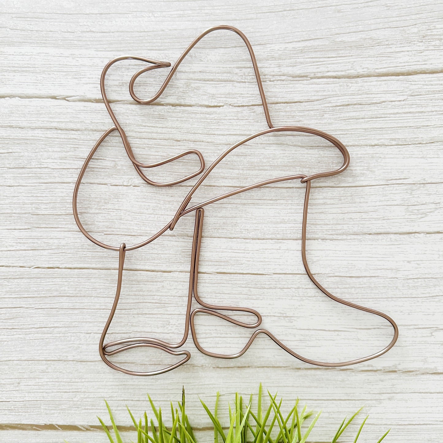 Handcrafted wire art of cowgirl boots and hat in mocha wire, hanging on a white wall with neutral western decor.