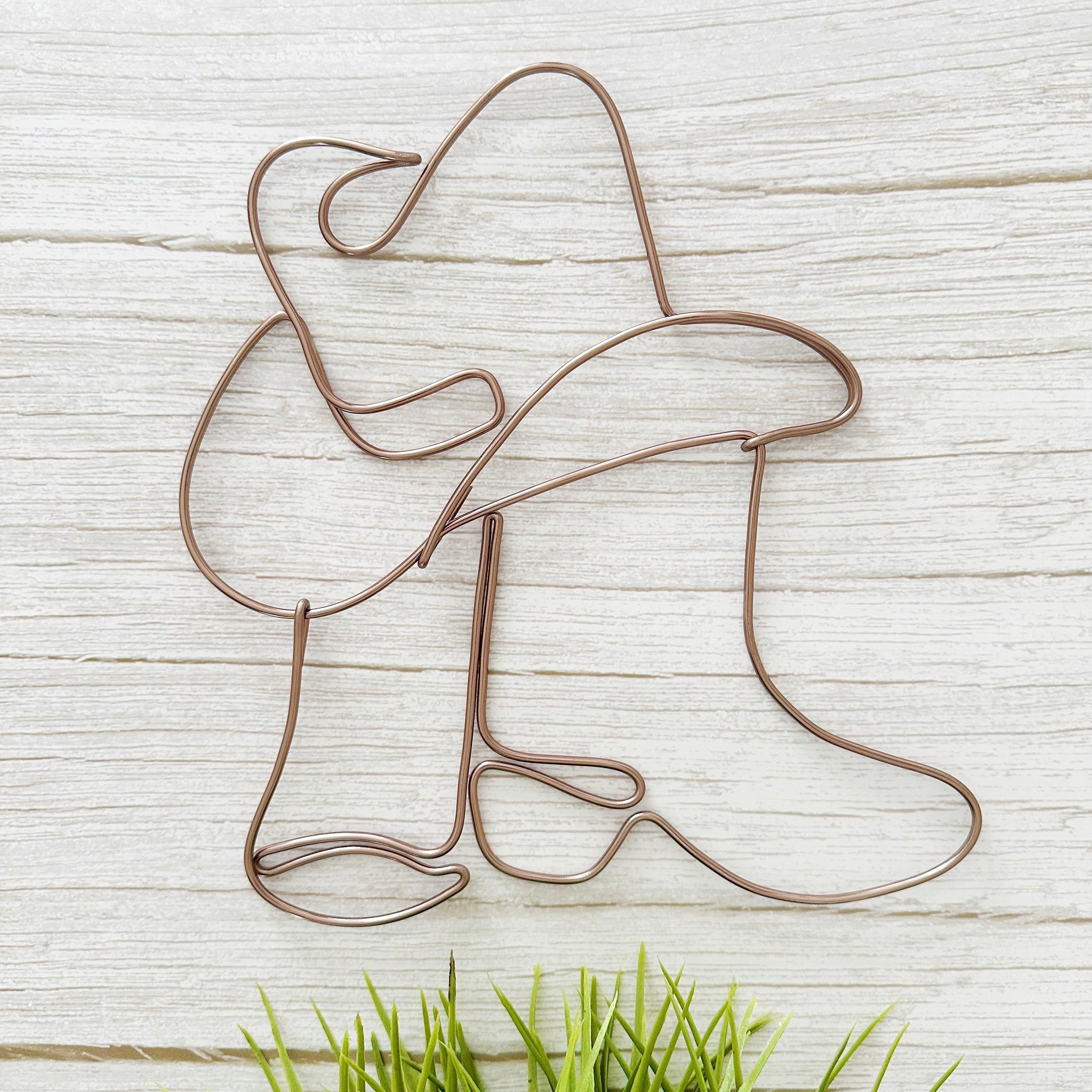 Handcrafted wire art of cowgirl boots and hat in mocha wire, hanging on a white wall with neutral western decor.