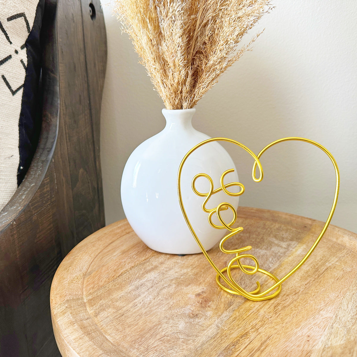 Gold wire heart-shaped decor on a wooden surface with a white vase and dried plants in the background.
