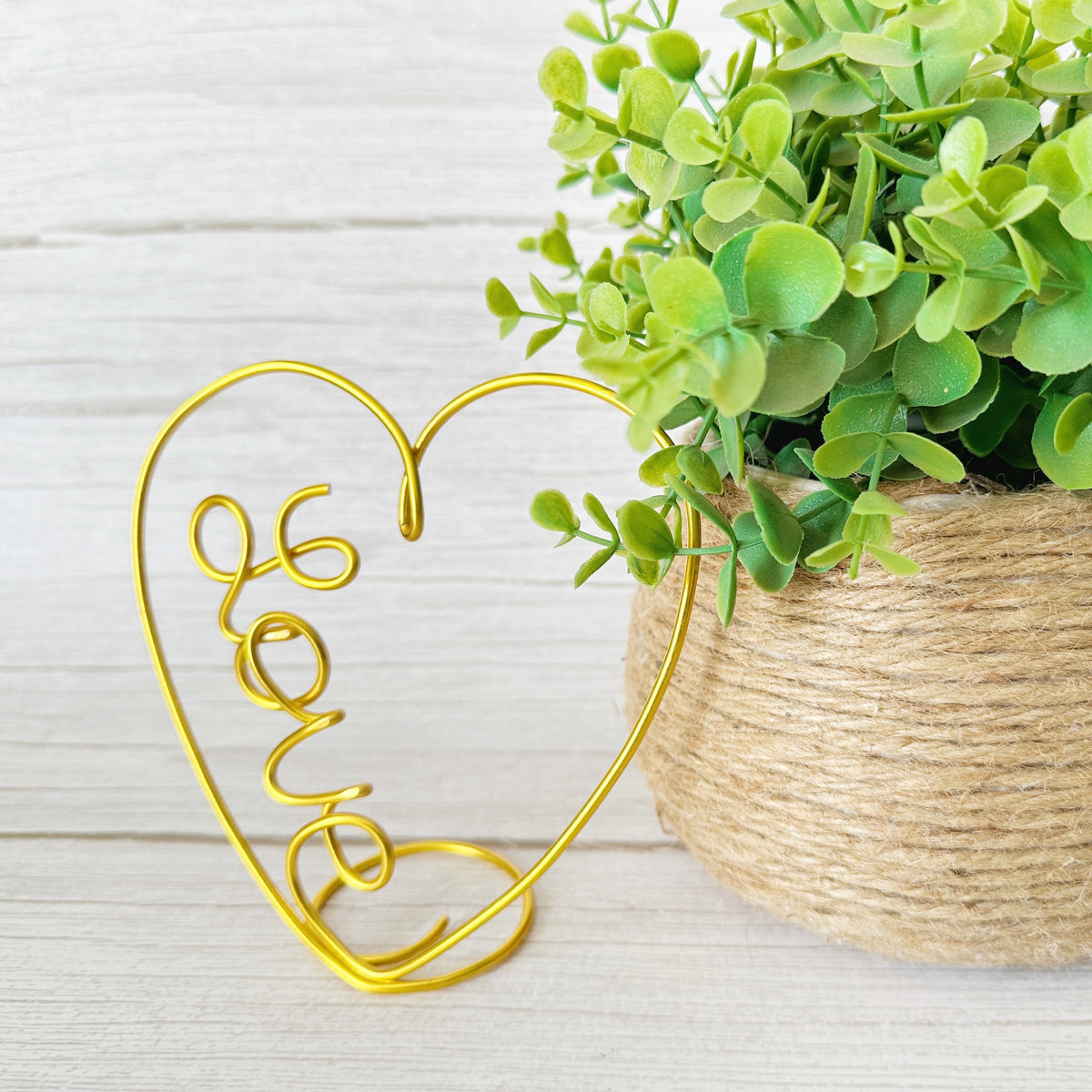 Gold wire heart-shape decor with love wire word inside sitting next to a faux plant on a light wooden surface