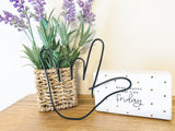 Decorative plant with lavender in a woven basket on a wooden surface, with a hand in the shape of ASL I love you made from black colored wire.