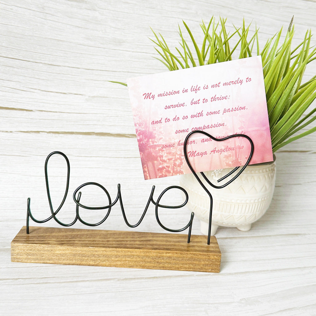 Decorative wire art sign with 'love' and heart design on a wooden base that can hold a photo or a note card. Faux plant in the background.