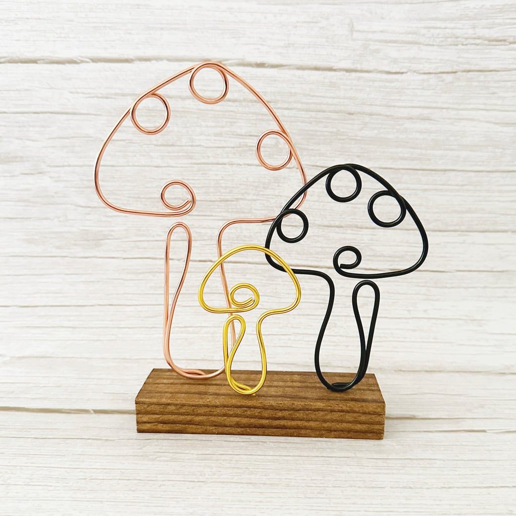 Handmade wire art mushrooms in copper, black, and gold on a rustic wood base, styled as whimsical home or shelf decor.