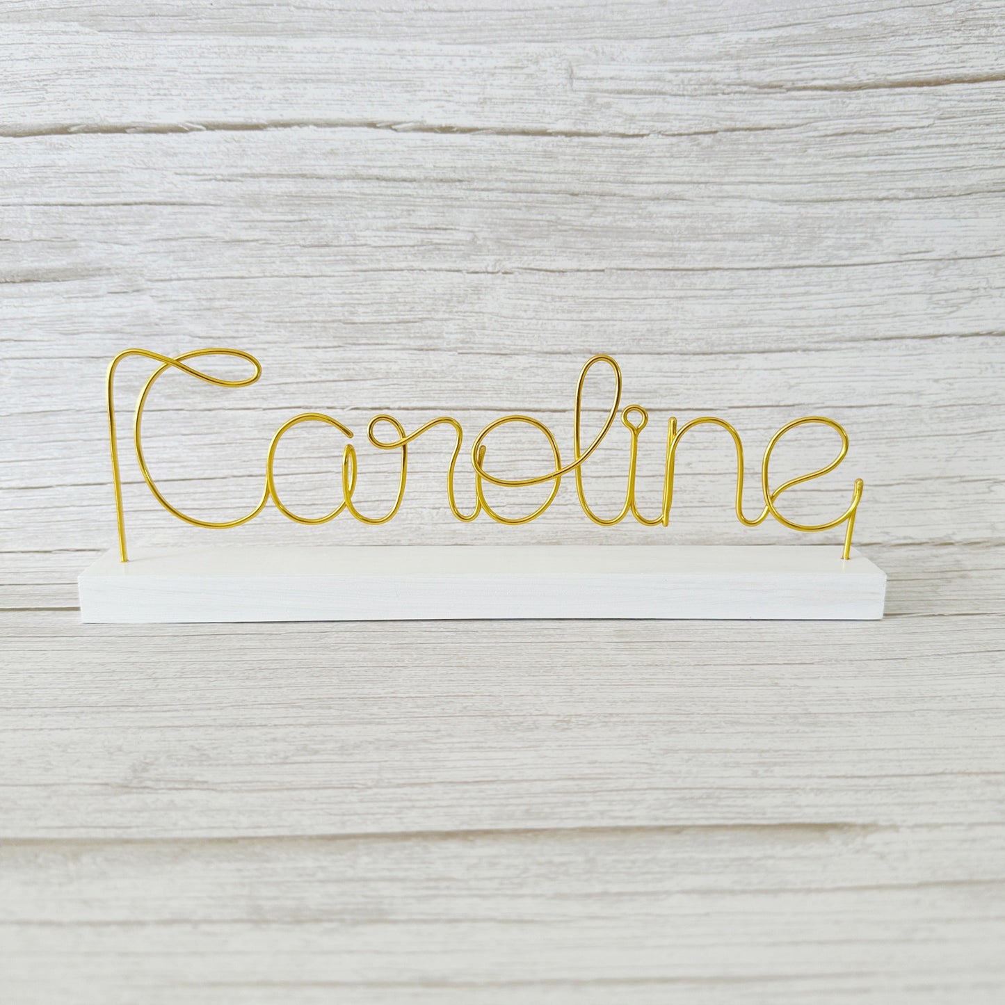 Gold wire name 'Caroline' on a white base against a light wooden background