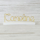 Gold wire name 'Caroline' on a white base against a light wooden background