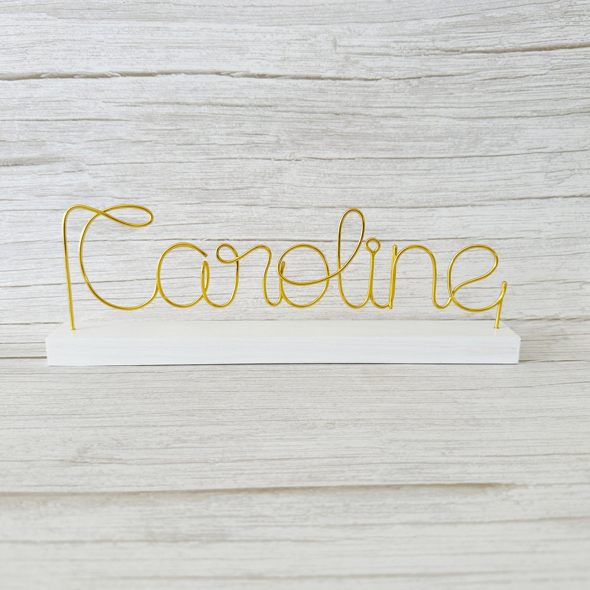 Gold wire name 'Caroline' on a white base against a light wooden background