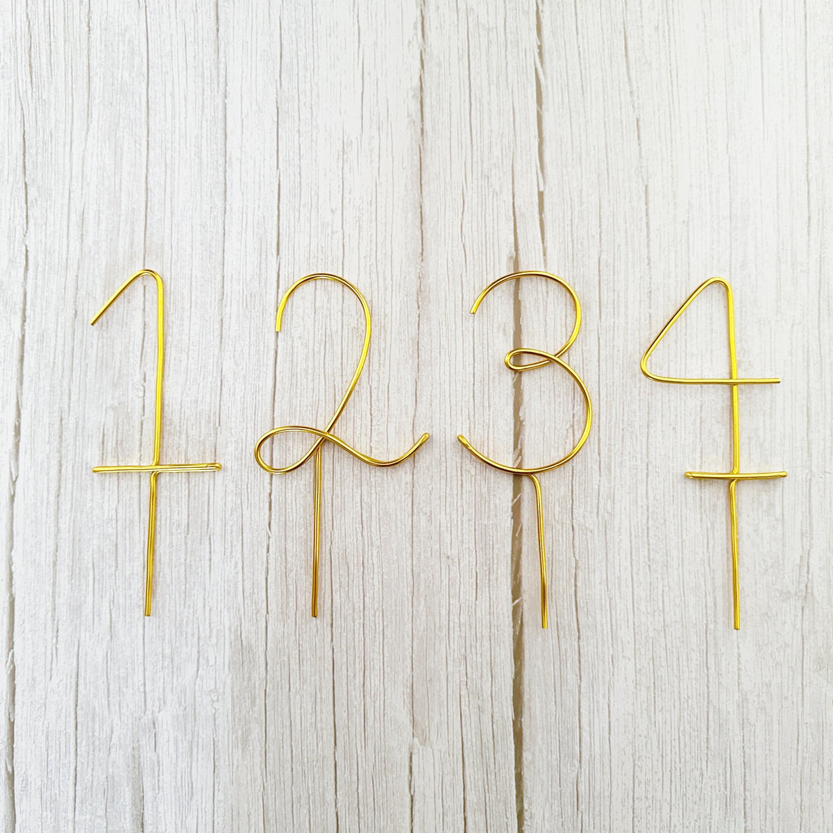 Handcrafted gold wire numbers 1, 2, 3, and 4 cake toppers with pick bases, displayed flat on a white wood background.