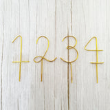 Handcrafted gold wire numbers 1, 2, 3, and 4 cake toppers with pick bases, displayed flat on a white wood background.