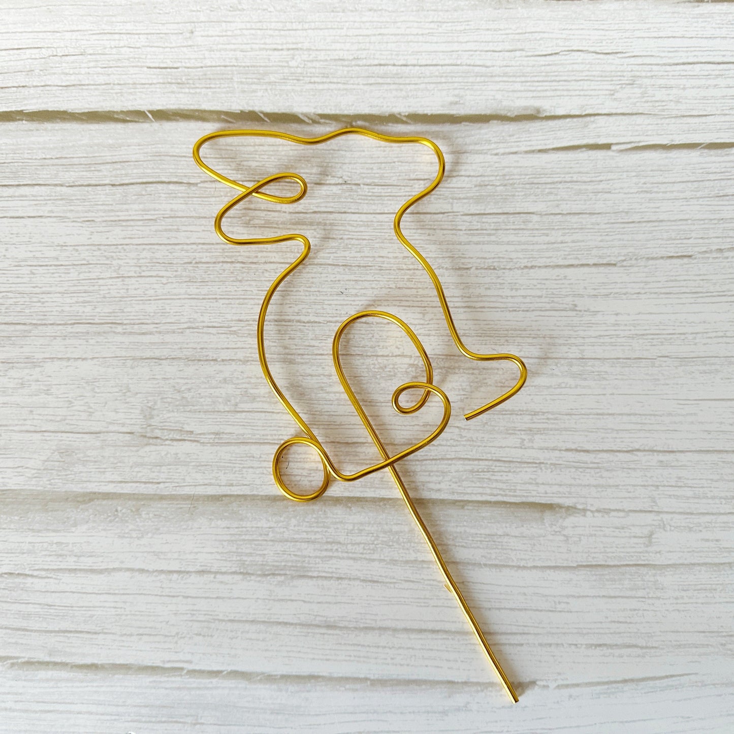 Gold wire sculpture of a bunny with a long stake laying on a wooden surface