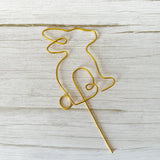 Gold wire sculpture of a bunny with a long stake laying on a wooden surface