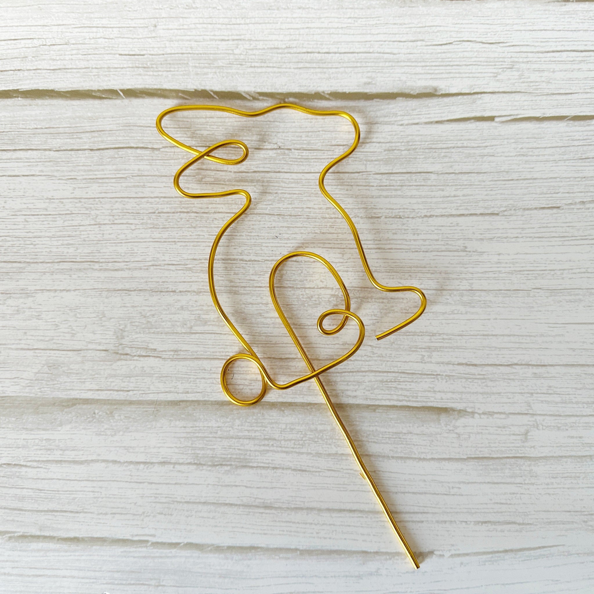 Gold wire sculpture of a bunny with a long stake laying on a wooden surface
