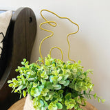 Gold wire bunny plant stake inserted into a faux decorative plant with green leaves on a wood table.