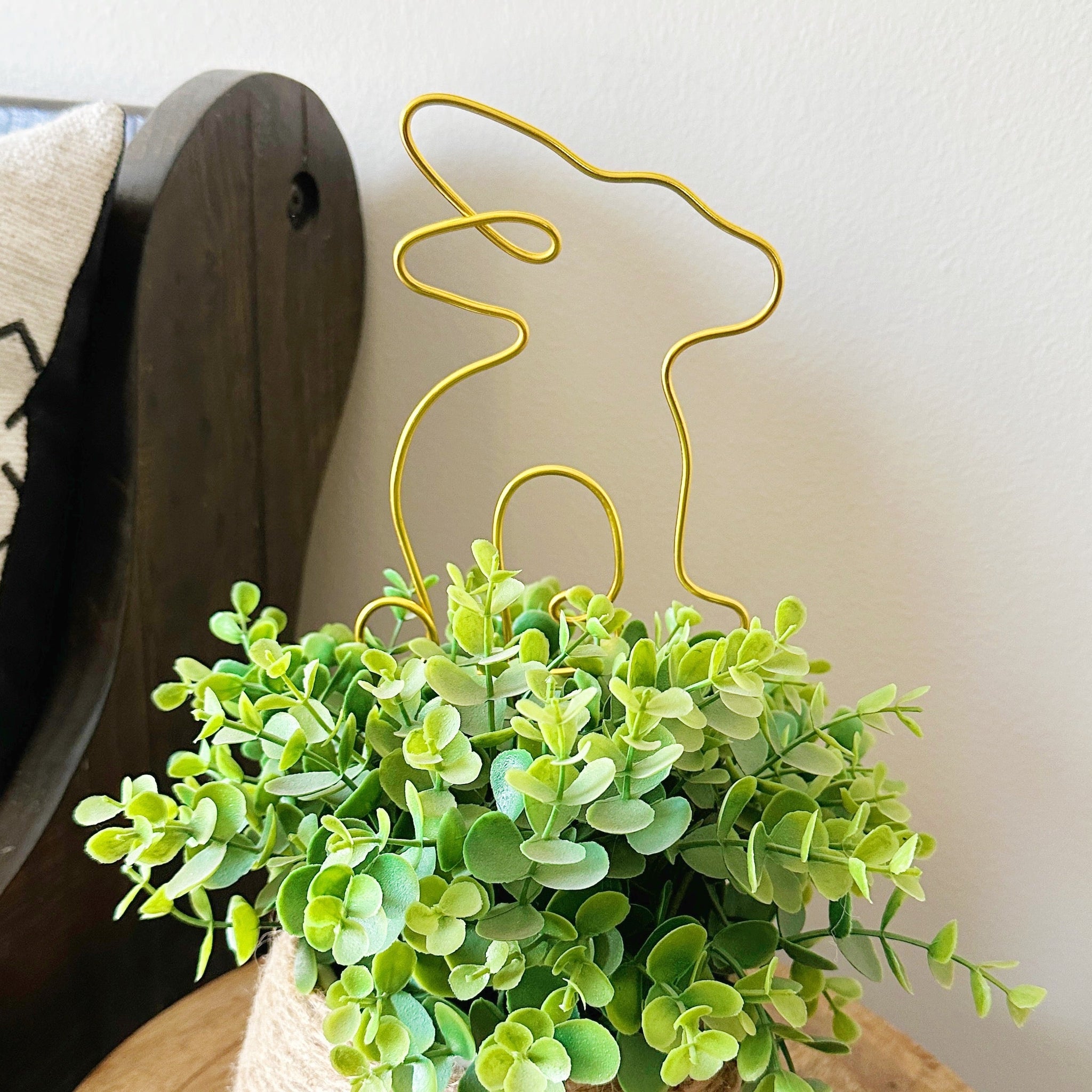 Gold wire bunny plant stake inserted into a faux decorative plant with green leaves on a wood table.