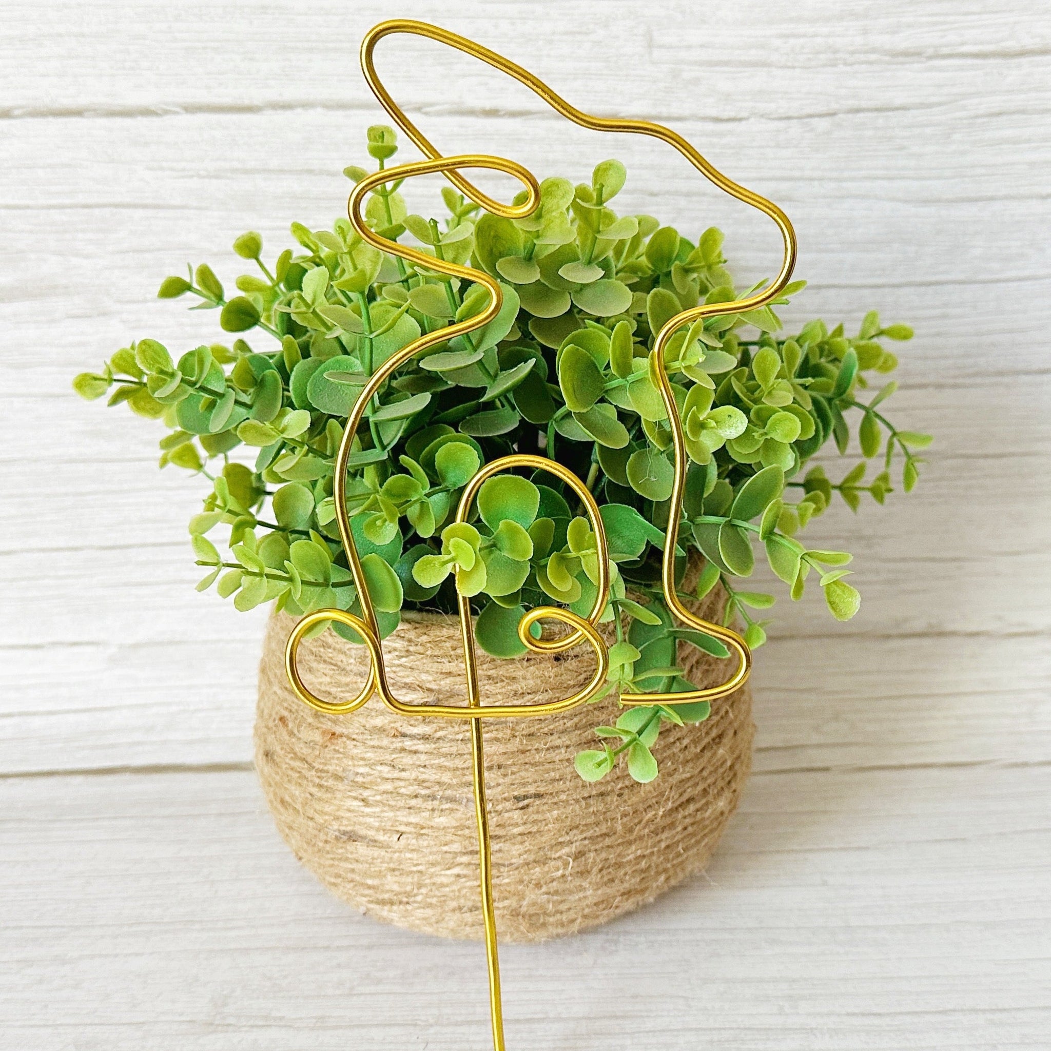 Gold wire bunny plant stake leaning against a faux decorative plant with green leaves on a light wood background.