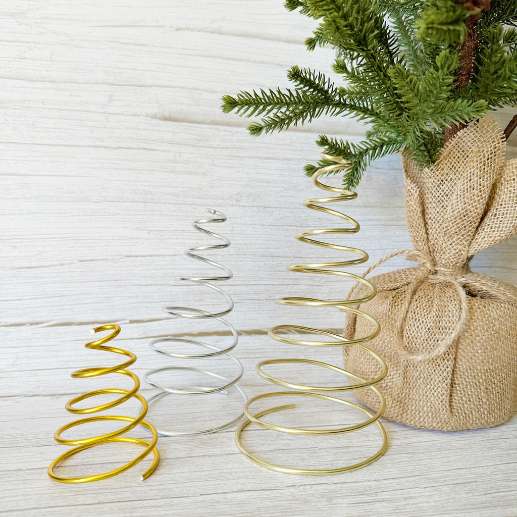 Three handcrafted wire spiral Christmas trees in gold and silver displayed on a light wood background in front of a burlap wrapped faux evergreen tree— minimalist handmade holiday décor by Emini Creations.