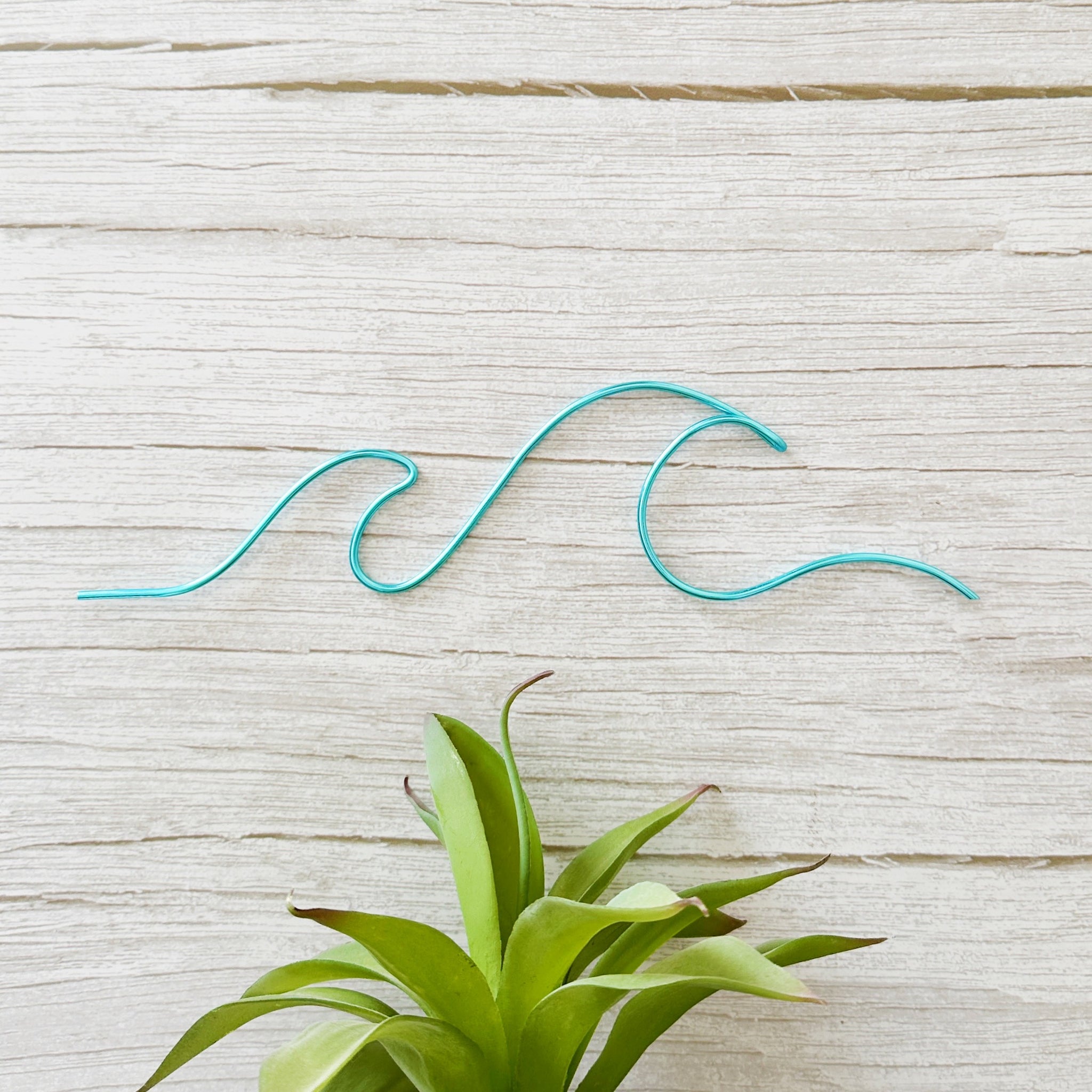 Handmade turquoise wave wire art displayed on a light wood background with green air plant – coastal-inspired minimalist decor