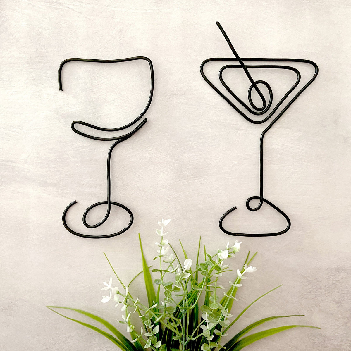 Two black wire art sculptures of a wine glass and a martini glass on a wall, with a faux underneath them.