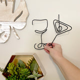 A hand posing with black wire art wine glass and martini glass against a beige wall along with succulents and a leaf wall decor.