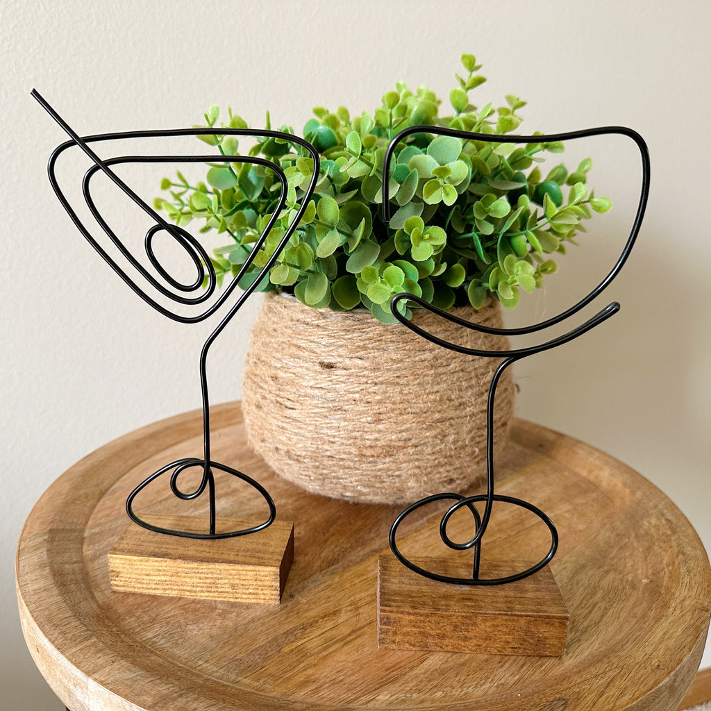 Decorative wire art shaped like a martini glass and a wine glass on a wooden stand with a faux plant in a boho vase in the background.