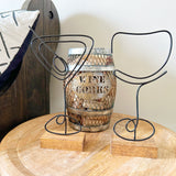 Decorative wine cork holder with a rustic wooden barrel design on a wooden surface with black wire martini and wine art sitting on a wooden table.