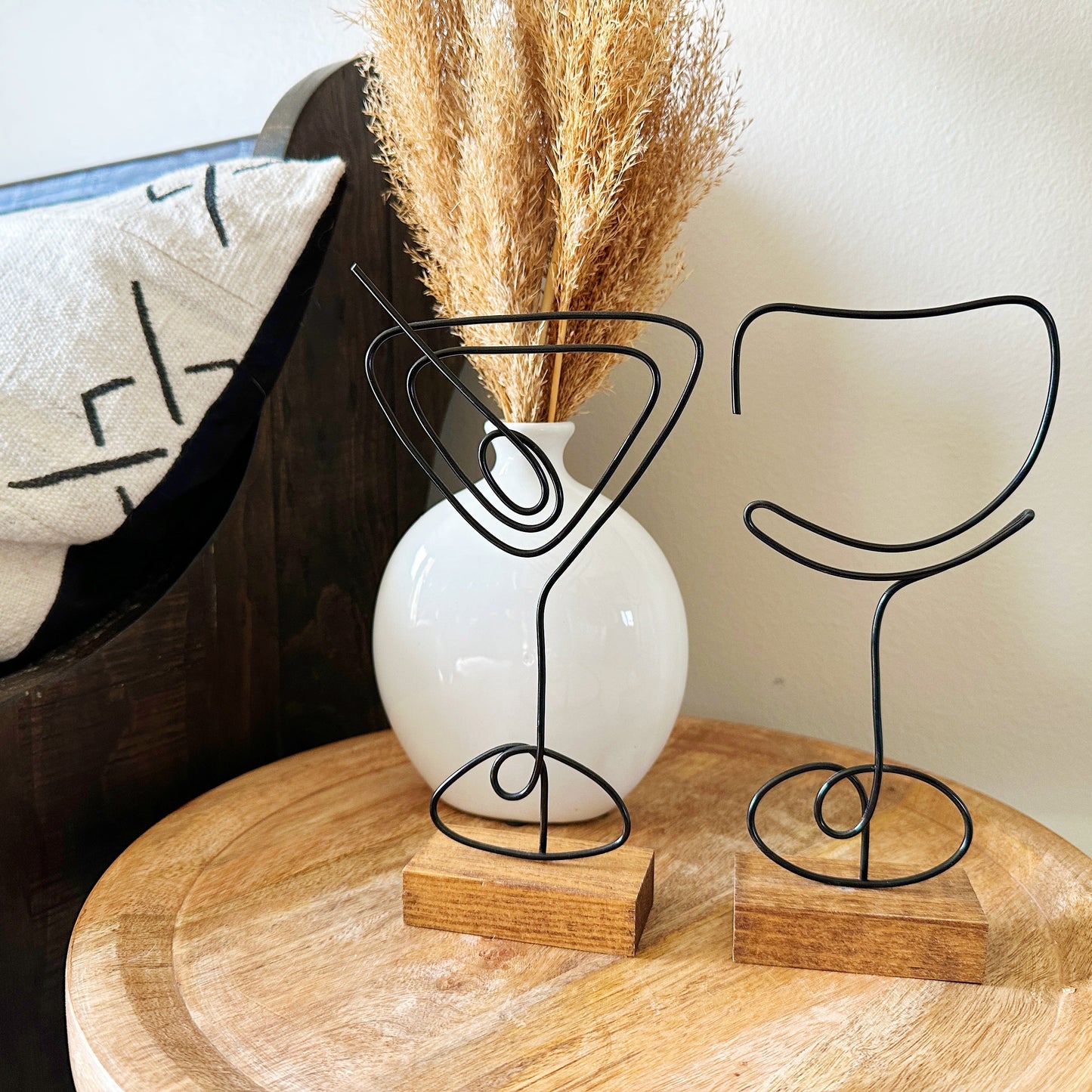 Decorative wire sculptures of a martini glass and a wine glass on a wooden surface with a white pillow in the background and a white vase with dried grass.