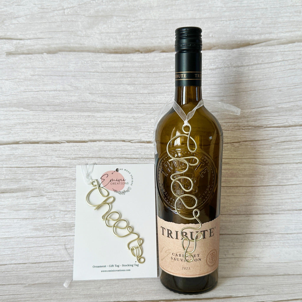 Handmade wire “cheers” tag tied around a wine bottle with white ribbon, shown with packaging from Emini Creations.