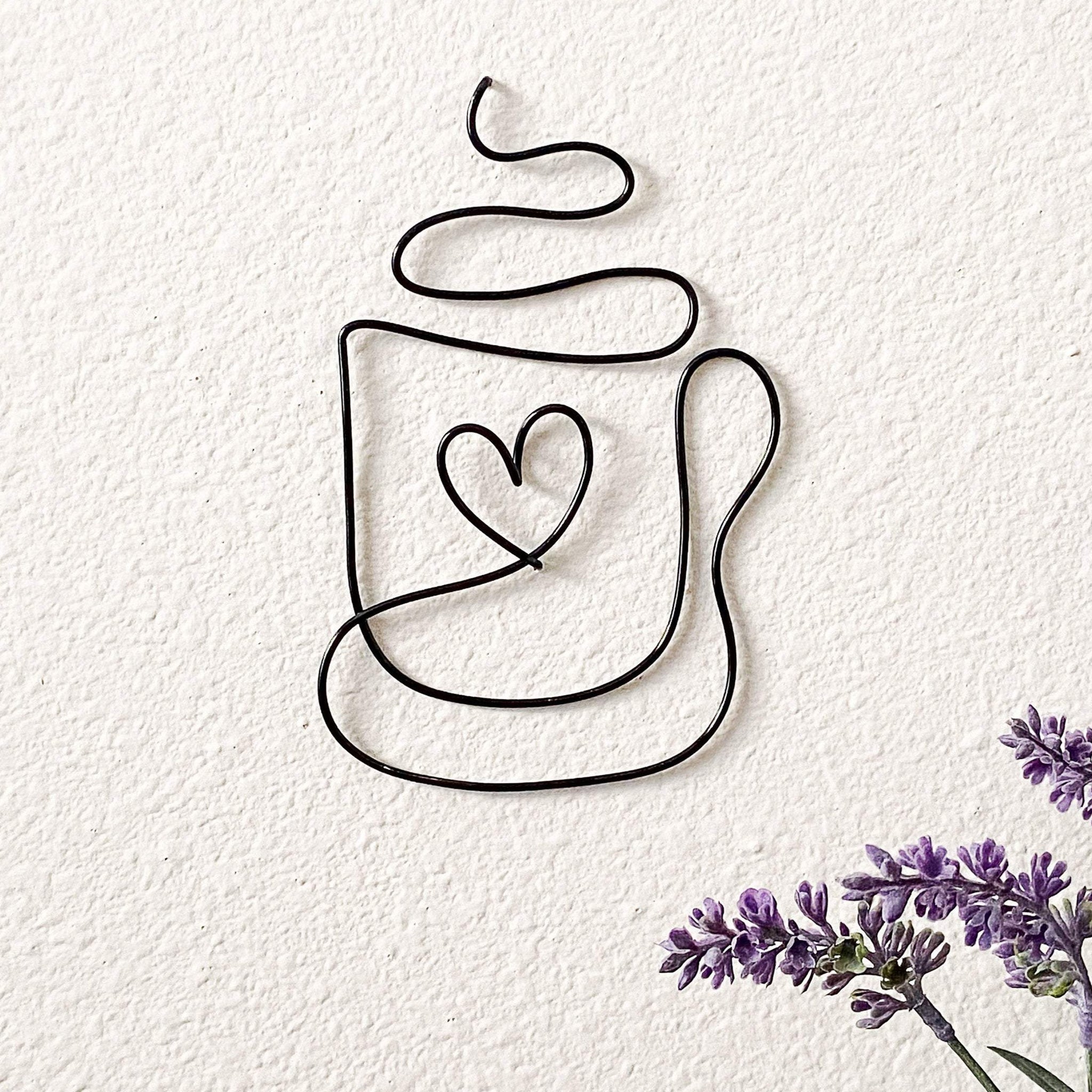 A wire coffee cup shaped wall decor with a heart inside the cup, mounted on a white wall with some lavender flowers beside it.